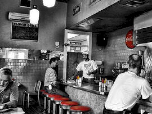 A diner with red stools and a checkered floor, featuring patrons at the counter and staff serving, in a retro black-and-white setting.