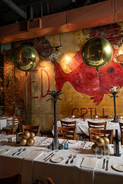 A dining room with a large red fish mural on the wall, gold balloons, and white tablecloths.
