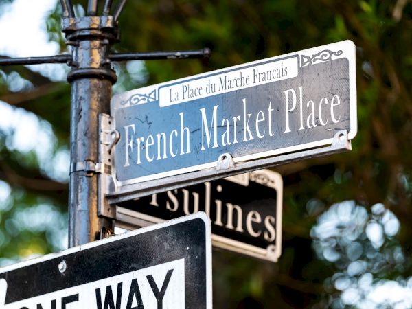A street sign for French Market Place and Ursulines Avenue with a one-way sign, surrounded by greenery.