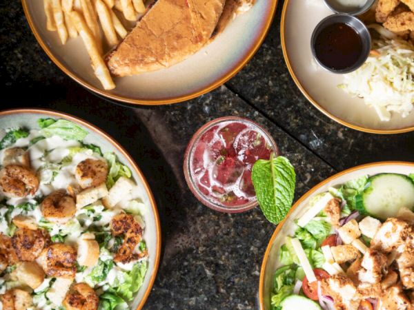 An overhead view of various dishes: a sandwich with fries, salads with toppings, a drink with mint, and a plate with dips.