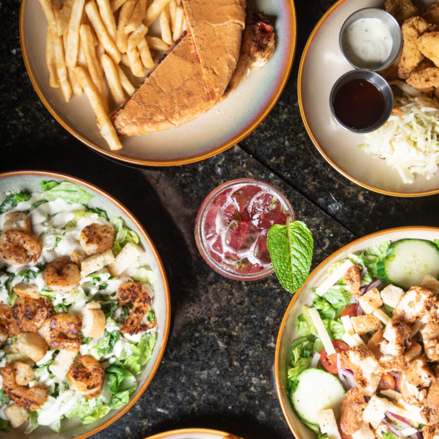 A selection of food on a table including salads, a sandwich with fries, and a drink.