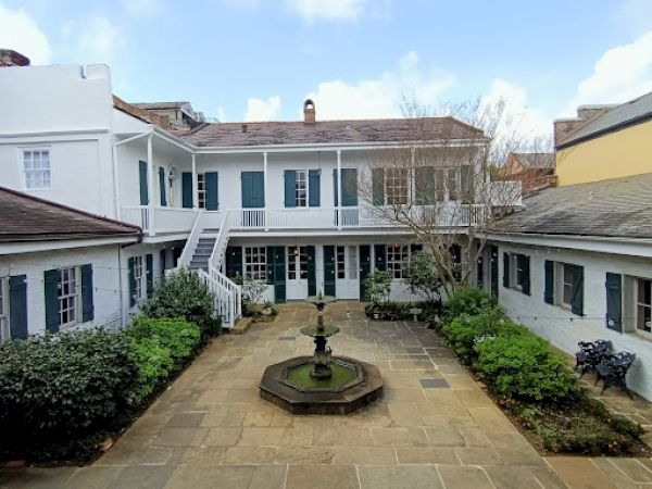 The image shows a courtyard with a central fountain, surrounded by white buildings with green shutters and a staircase leading to the upper level.