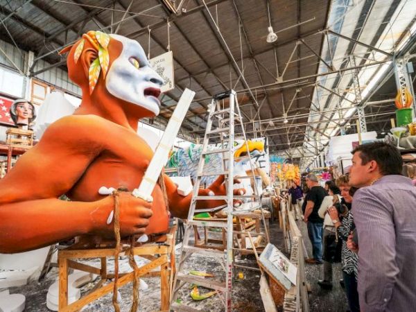 A workshop with giant orange statue heads under construction; workers sculpt with masks, ladders, and paint, while visitors watch in an art market.