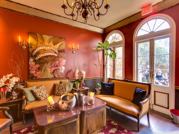 A cozy, stylish lounge with red walls, antique furniture, and warm lighting; a sofa, coffee table, plants, artwork, and arched windows.