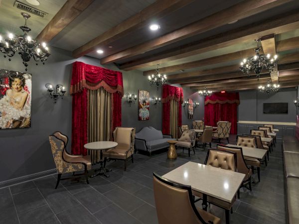 A stylish, dimly lit lounge with red curtains, chandeliers, leather chairs, small tables, and framed art along dark walls, arranged for intimate seating.