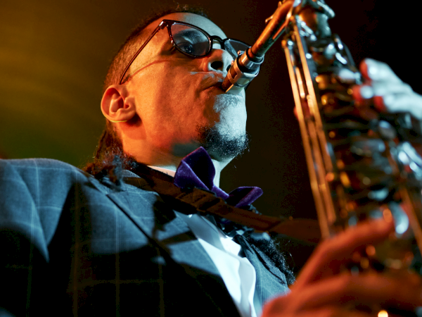 A saxophonist in a suit and sunglasses plays a brass instrument on stage, under warm concert lighting, intense and focused.