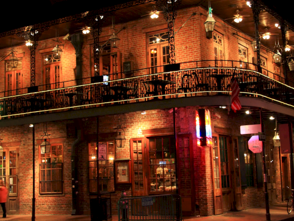 A two-story brick building at night, wrought-iron balcony, warm lights, lanterns, street corner cafe vibe with signage and pedestrians.