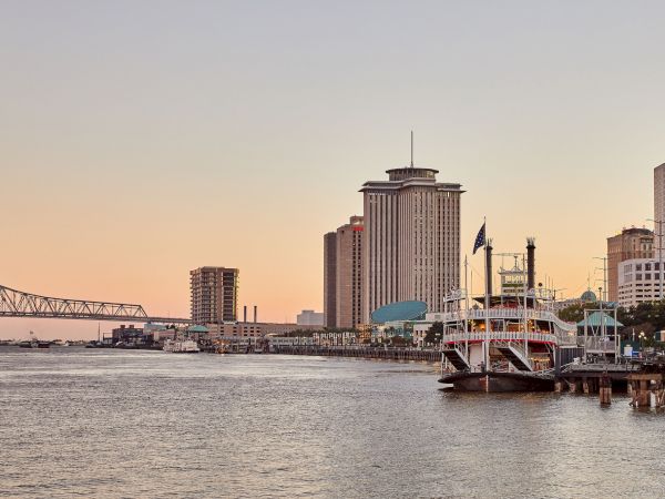 A riverfront skyline with a tall downtown tower, a docked cruise ship with flags, and a bridge stretching across the water at dusk.