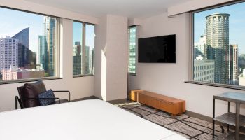 A modern hotel room with a bed, chair, TV, and city skyline view through large windows on two walls.