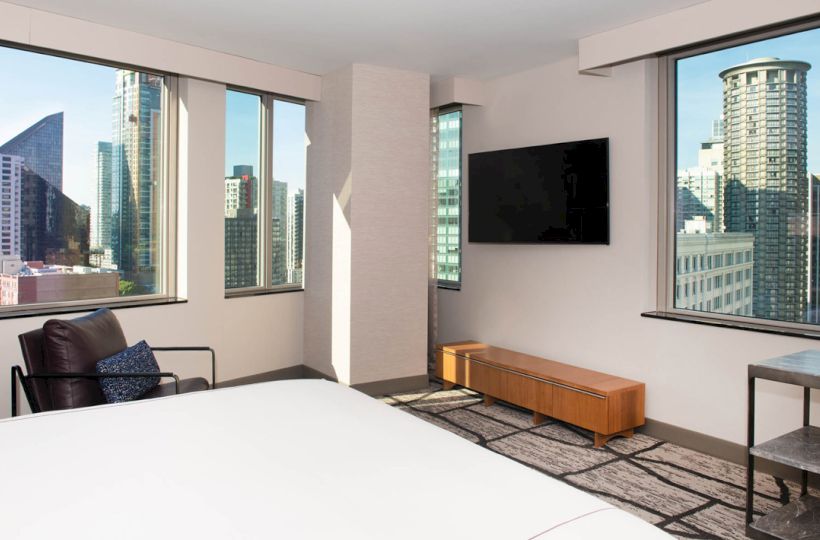 A modern hotel room with a bed, chair, TV, and city skyline view through large windows on two walls.