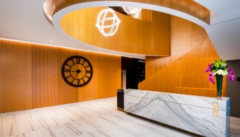 The image shows a modern lobby with a marble reception desk, wooden accents, a large wall clock, and decorative ceiling lights.