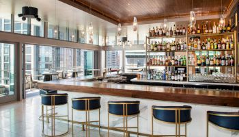 A modern bar interior with a wooden counter, stylish barstools, and shelves stocked with a variety of bottles. Large windows offer a view.