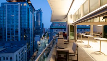 The image shows a modern balcony with seating overlooking a cityscape at dusk, featuring tall buildings and illuminated windows.