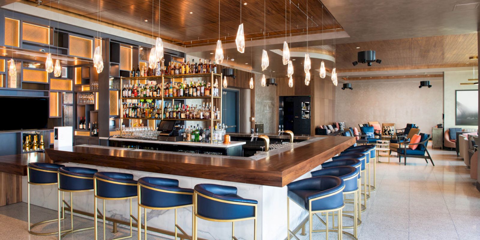 The image shows a sleek, modern bar with blue stools, pendant lights, and a fully stocked back wall of bottles.