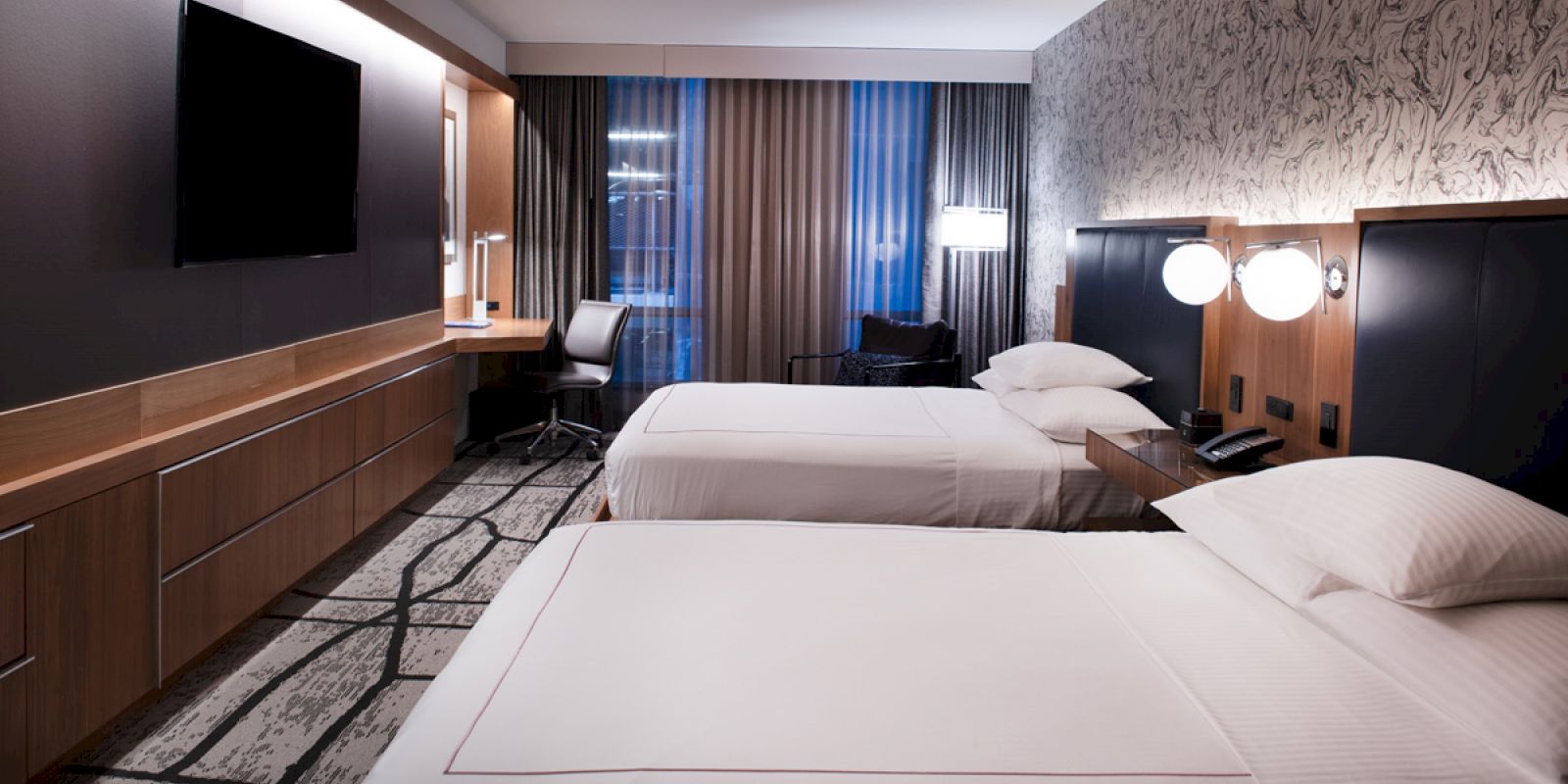 The image shows a modern hotel room with two beds, a large TV, desk, chair, and patterned walls and carpet.