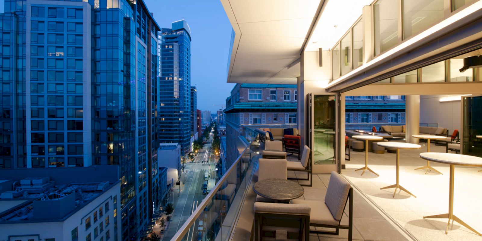 A modern rooftop balcony with seating overlooks a city street lined with tall buildings, under a dusky blue sky.