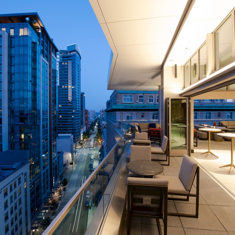 A modern urban balcony with seating overlooks a city street flanked by tall buildings, captured during the evening.