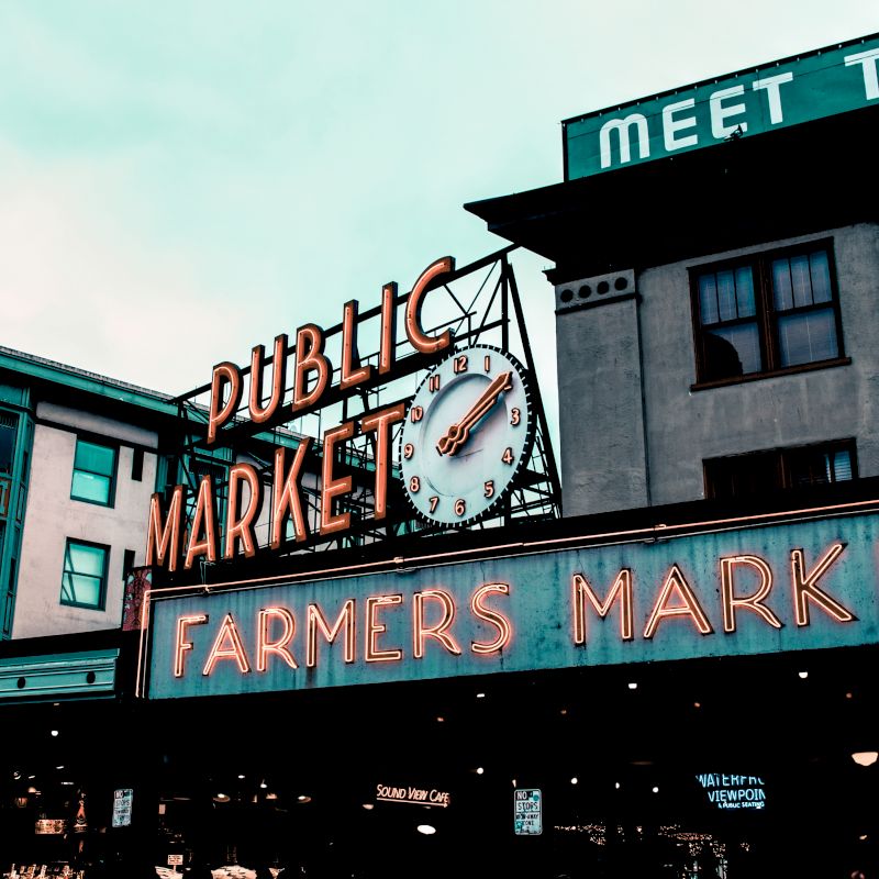 A neon sign reads “Public Market” and “Farmers Market” with a clock face in between, set against historic buildings under a cloudy sky.