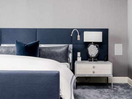 A modern bedroom features a navy bed with white and blue bedding, a sleek nightstand, and a lamp, all on a plush carpeted floor.