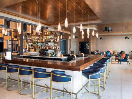 A modern bar with blue chairs, a wooden counter, pendant lights, and shelves stocked with bottles, in a stylish lounge setting.