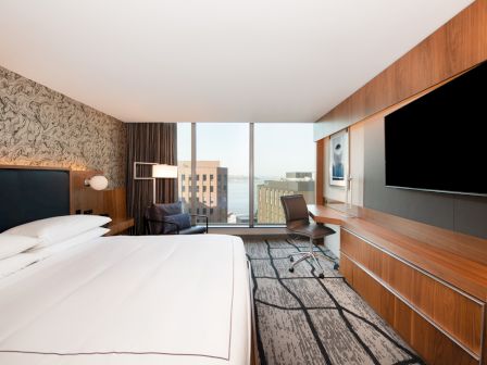 A modern hotel room with a large bed, wooden furniture, a desk, and a city view through large windows.