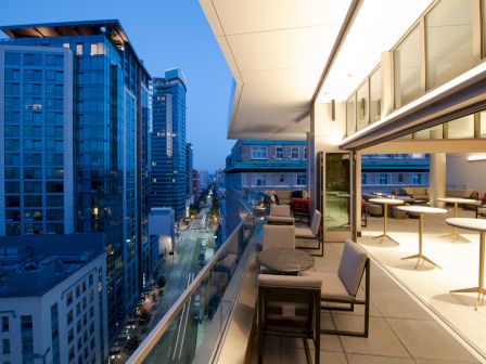 A modern rooftop terrace with tables and seating overlooks a cityscape at dusk. Vibrant city lights illuminate the surrounding high-rises.