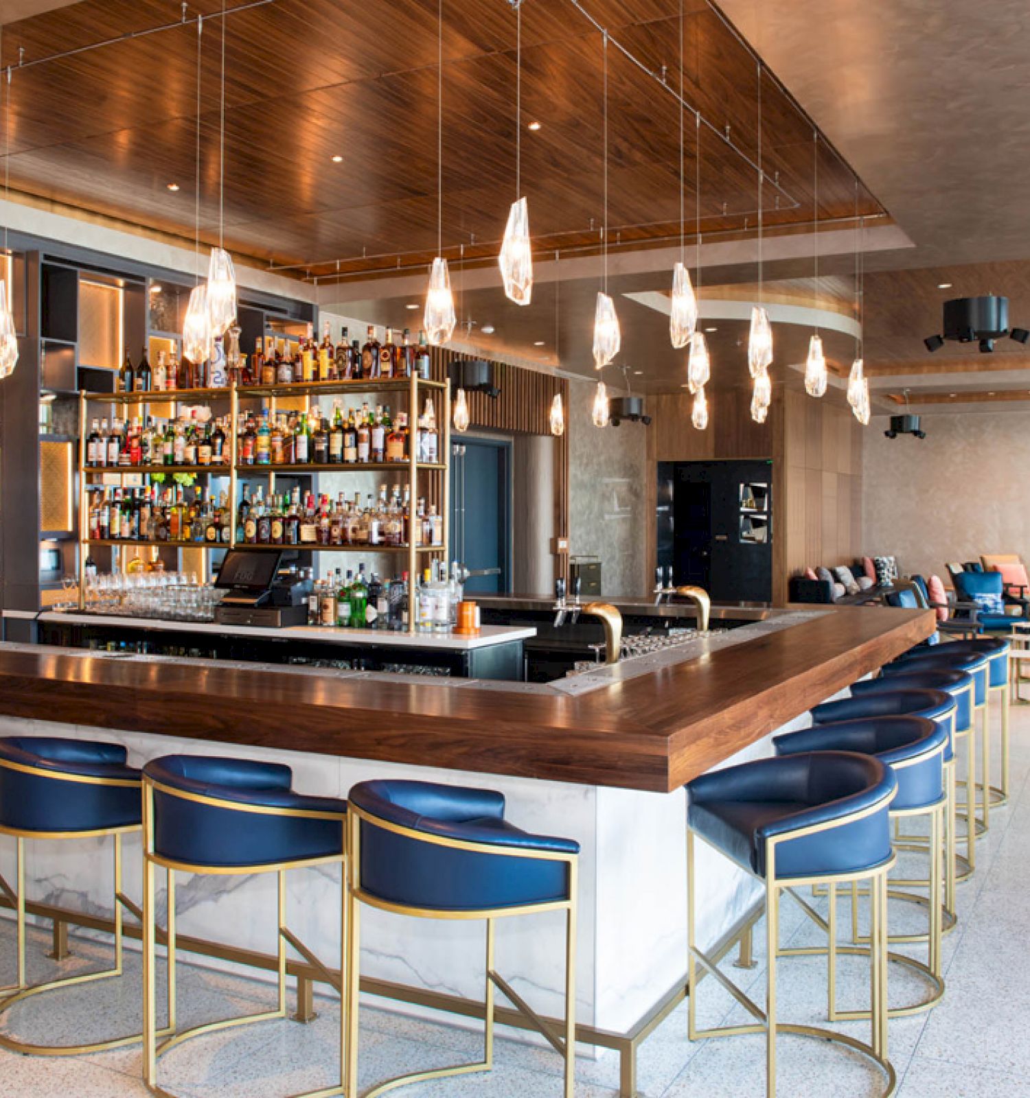 A modern bar with blue cushioned stools, a well-stocked liquor selection, pendant lights, and cozy seating in the background.