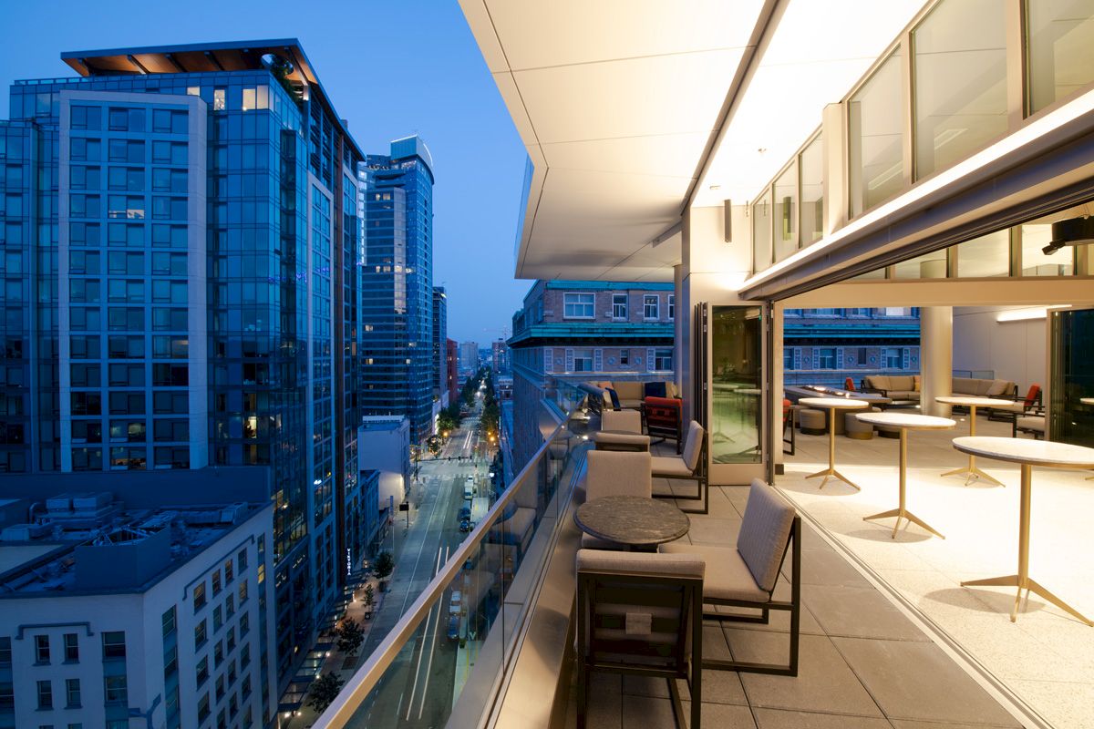 A modern urban rooftop terrace with tables and chairs overlooking a city street and high-rise buildings at dusk.