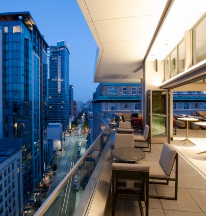 A modern balcony scene at dusk, overlooking a cityscape with tall buildings, featuring outdoor seating and a contemporary design.