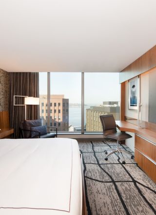 A modern hotel room with a large bed, desk, chair, wide TV, and a view of buildings and water through large windows.