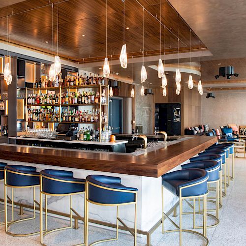 A modern bar with a sleek design, featuring blue chairs, a stocked bar, and ambient lighting, creating a chic and inviting atmosphere.