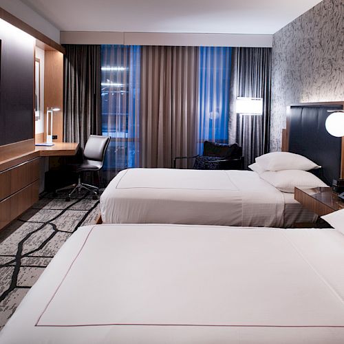 The image shows a hotel room with two beds, a TV, a work desk, and modern decor. There are patterned curtains and a stylish wall design.