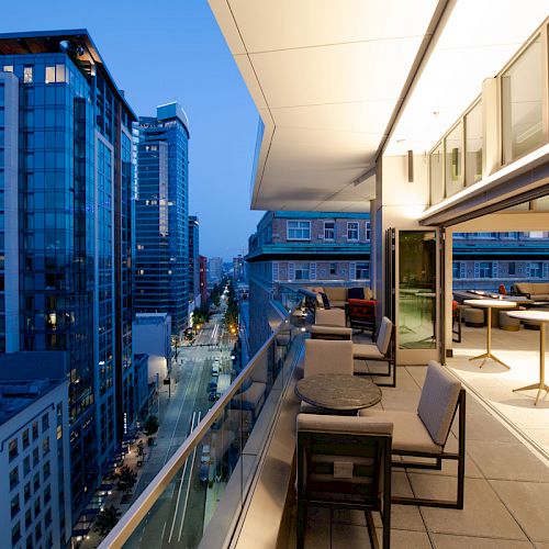 Cityscape view from a modern balcony at dusk, featuring a glass-enclosed area with tables and chairs, overlooking urban buildings.