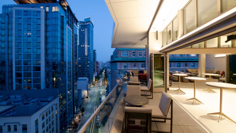 A modern urban balcony at dusk, overlooking a city street with tall buildings and a cozy seating area under warm lights.