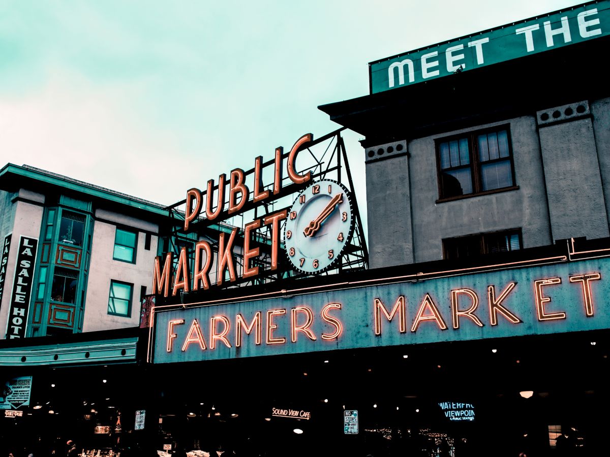 The image shows the iconic neon signs for a Public Market and Farmers Market with a clock on a building, possibly Pike Place Market.