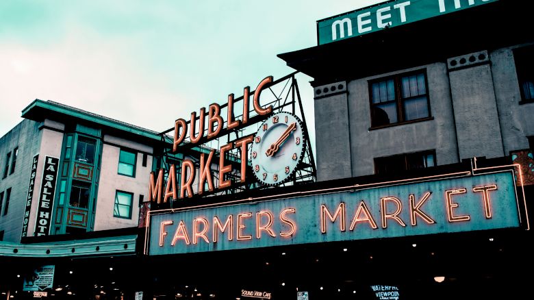 The image shows the iconic neon signs for a Public Market and Farmers Market with a clock on a building, possibly Pike Place Market.