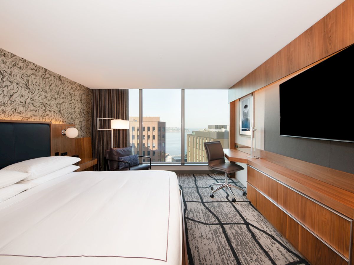 A modern hotel room with a large bed, desk, and wall-mounted TV, offering a city view through large windows.