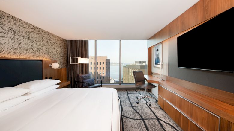A modern hotel room with a large bed, desk, and wall-mounted TV, offering a city view through large windows.