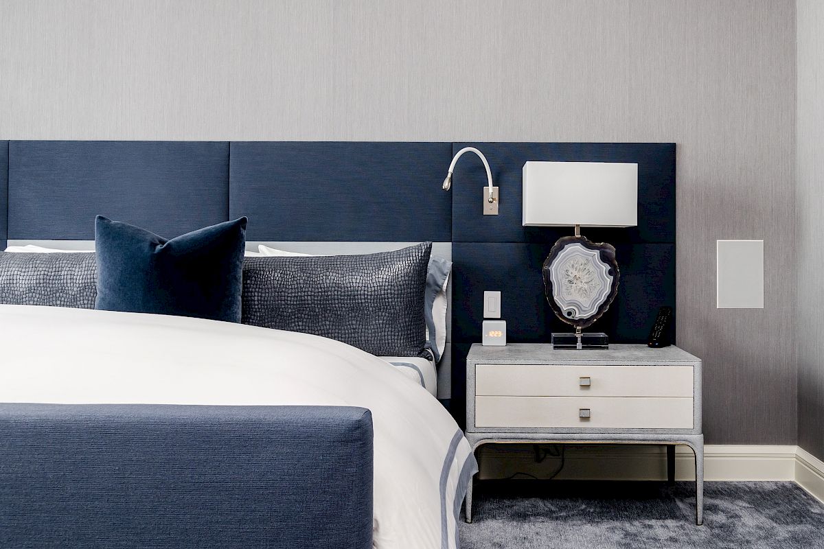 A modern bedroom with a blue headboard, white bedding, and a nightstand holding a lamp and decor items against a grey wall.
