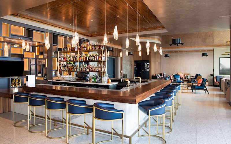 A modern bar with blue stools, a well-stocked liquor shelf, hanging lights, and a spacious lounge area with seating.