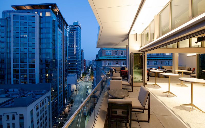 A modern rooftop terrace at dusk, featuring outdoor seating and tables, overlooks a city street lined with tall buildings.